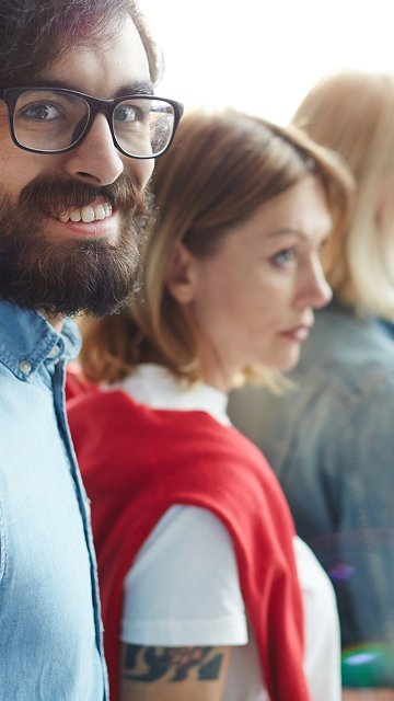 A bearded man in glasses standing in line, smiling, attention forward — paying attention on someone's behalf